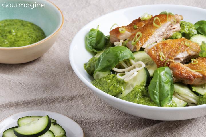 A white bowl with noodles, chicken and cucumber next to a bowl with green sauce and a plate of cut cucumbers