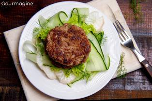 A white plate with a bed of cucumber salad topped with burger patties with Chipeño
