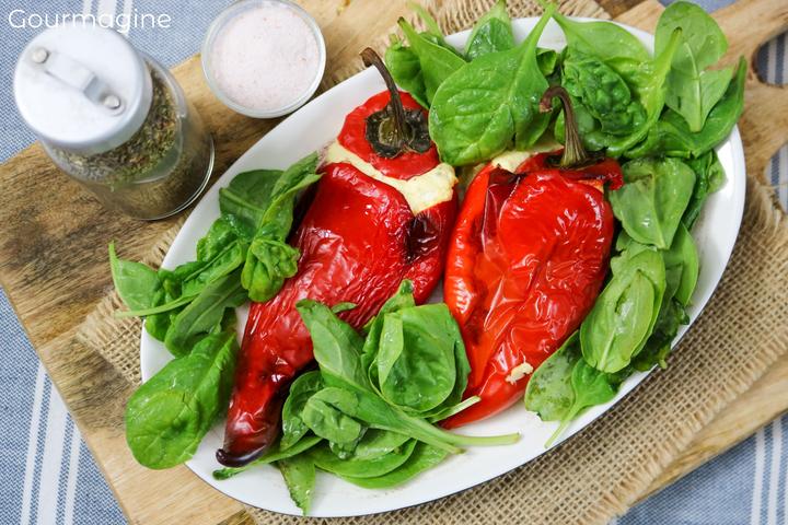 A white plate with two red peppers stuffed with white cheese and arranged on a bed of spinach salad next to a glass container with oregano