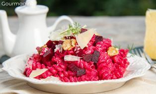 A white plate with pink pasta, beetroot pieces, cheese splinters and walnuts on a white tablecloth and a white teapot in the bacckground
