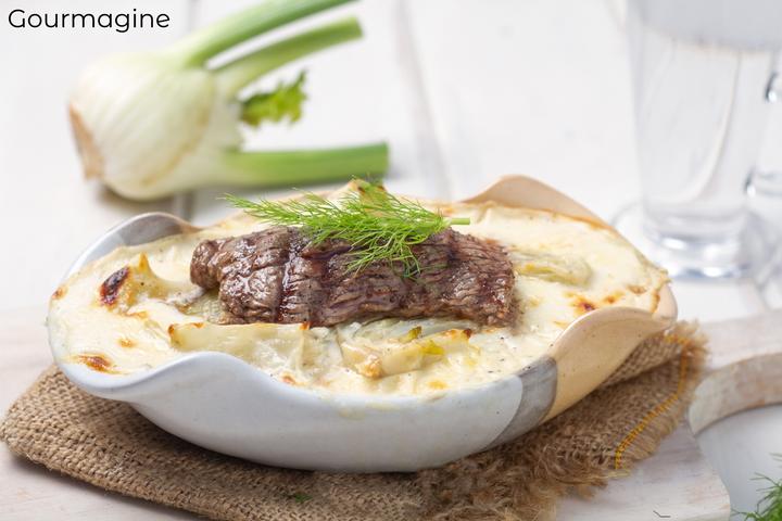 A white baking dish with a fennel gratin topped with a beef steak on a wooden board and with a fennel in the background