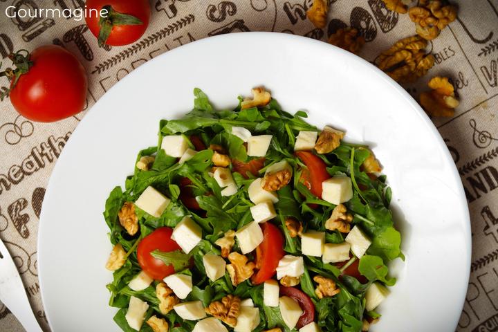 A white plate with green rocket salad, red cherry tomatoes, white mozzarella pieces and brown walnuts on a grey table cloth with tomatoes and walnuts in the background