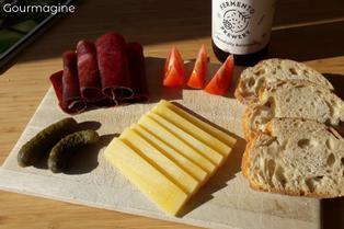 Wooden board with cut yellow mountain cheese, rolls of beef, tomato wedges and slices of bread next to a craft beer