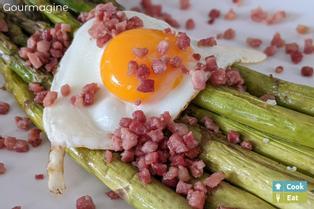 Green asparagus on a whtie plate covered with bacon cubes and a fried egg