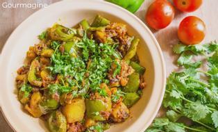 Ein weisser Teller gefüllt mit angebratenen grünen Peperoni, Kartoffeln und Zwiebeln an einer Curry-Sauce auf einem Tisch mit Tomaten und Koriander