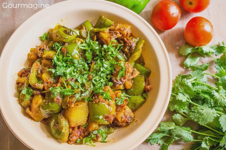 Ein weisser Teller gefüllt mit angebratenen grünen Peperoni, Kartoffeln und Zwiebeln an einer Curry-Sauce auf einem Tisch mit Tomaten und Koriander