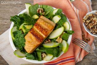 White plate with green rocket, apple slices and a salmon piece next to a bowl with nuts