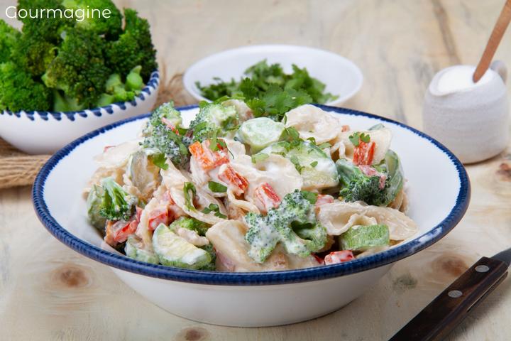 A white bowl filled with farfalle, broccoli, peppers and courgettes covered with a salad dressing